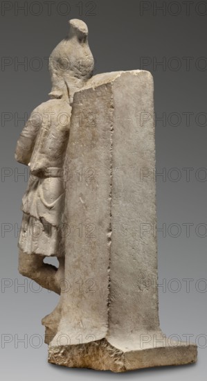 Fragment of a monopodium, unknown Roman workshop, Roman sculpture of Attys myth., 2nd century, Roman Empire 27 BC–476, sculpture; height 77 cm, width 31 cm, depth 34 cm, weight 64 kg., A pale stone sculpture viewed from an angle showing the back and side of a standing figure partially obscured by a tall rectangular slab; the figure wears a short garment with folds and a belt and has one leg bent with a foot resting on a base. The stone surfaces show rough and smoother textures with weathered edges and tool marks. Visible colors include beige, off-white, light gray, tan, and hints of brown.