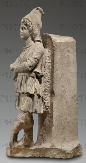 Fragment of a monopodium, unknown Roman workshop; Roman sculpture depicting Attys myth.; 2nd century, Roman Empire 27 BC–476; sculpture; materials/technique unspecified; height 77 cm, width 31 cm, depth 34 cm, weight 64 kg., A stone sculpture of a standing figure leaning against a rectangular block, shown in profile facing left, with a textured surface in beige and gray tones. The figure wears a tall, rounded headdress and a layered tunic with visible folds and drapery; the arms are crossed at the chest and the legs are slightly bent with one foot placed forward. The rectangular support has a rough, pitted side adjacent to the figure and a smoother front, both sharing the same stone color palette of pale beige and light gray. The base is irregular and rough, matching the muted beige-gray of the sculpture, and subtle shadows emphasize the carved details and contours.