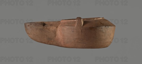 Lamp with a tragic mask, unknown Italian workshop. Oil lamp, single-burner, Roman, Buchi type IX-b = Loeschcke type IXb, probably 2nd half 1st–beginning 2nd century. From a mold. Round vessel with arms drooping to the outer, separated from the disc by a convex edge; two bars arranged symmetrically on the arms. Concave, flat disc with two olive holes located on either side of a convex theatrical tragic mask. Medium-length wick channel decorated with a central groove and a small ventilation hole in the groove; rounded end of the burner. Slight trace of use dark discoloration near the wick hole. On the bottom, a convex inscription workshop trademark: COMMUNIS. Height c. 9 cm, width c. 4.4 cm, depth c. 3.3 cm., A single elongated object resembling a carved vessel or model boat fills the center of the image, shown in profile against a plain gray background. The object has an overall earthy brown and reddish-brown surface with areas of darker brown and muted orange tones, and a few lighter b...