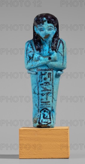 Queen Maatkare's shabti, Divine Wife of Amun, cartouches, hieroglyphs, uraeus iconographic, Maatkare ca. 1069–945 BC, wigs, funerary figurines, grave goods, mummy-shaped figurine. Arms crossed on the chest, right folded over left. Hoes in each hand. On the back is a basket slung on strings over both shoulders. Facial details, wig, and tools painted in a dark color. Three-part smooth wig. Uraeus above the forehead. Bracelets on the wrists. Painted inscription on the front of the legs in one wide column framed with lines: Shd Osiris, the wife of the god, the lord of both lands, Maat-ka-re. Third Intermediate Period; 21st Dynasty, Egypt. Shabti, height 12 cm, width 4.4 cm., A small blue figurine of a standing human figure with crossed arms, mounted on a rectangular tan base, shown against a plain light gray background. The figure is predominantly bright turquoise blue with darker blue to black coloration on the hair, shoulders, and facial features, and some darker blue markings on the tor...