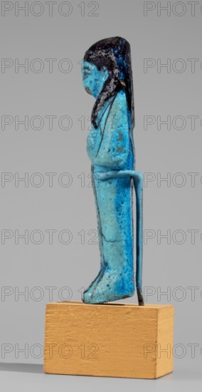 Queen Maatkare's shabti, Divine Wife of Amun, cartouches, hieroglyphs, uraeus iconography, Maatkare ca. 1069–945 BC, wigs, funerary figurine, grave good, mummy-shaped figurine. Arms crossed on the chest, right folded over left, hoes in each hand. On the back is a basket slung on strings over both shoulders. Facial details, wig, and tools painted in a dark color. Three-part smooth wig; a uraeus above the forehead; bracelets on the wrists. Painted inscription on the front of the legs in one wide column framed with lines: Shd Osiris, the wife of the god, the lord of both lands, Maat-ka-re. Third Intermediate Period, 21st Dynasty, Egypt. Shabti, height 12 cm, width 4.4 cm., A small standing figurine in shades of bright blue and turquoise with darker blue and black areas, shown in profile on a rectangular light brown wooden base; the figure has long dark hair, a rounded head and simple facial features, arms folded across the chest, a slim torso and long skirted lower body with subtle darker...