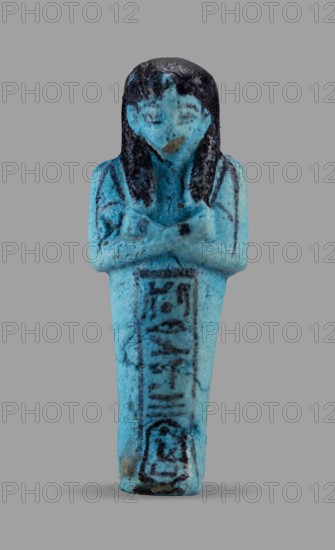Queen Maatkare's shabti, Divine Wife of Amun, cartouches, hieroglyphs, uraeus iconographic, Maatkare ca. 1069–945 BC, wigs, funerary figurines, grave goods, mummy-shaped figurine. Arms crossed on the chest, the right folded over the left, holding hoes in each hand. On the back is a basket slung on strings over both shoulders. Facial details, wig, and tools painted in a dark color. On the head is a three-part smooth wig with a uraeus above the forehead. Bracelets on the wrists. Painted inscription on the front of the legs in one wide column framed with lines reading: Shd Osiris, the wife of the god, the lord of both lands, Maat-ka-re. Third Intermediate Period, 21st Dynasty, Egypt; shabti, height 12 cm, width 4.4 cm., A small figurine in shades of bright turquoise and blue stands upright against a gray background, depicting a human form with folded arms and a long, tapered lower body; the head and hair are darker, nearly black, with visible streaks and textured surface, and there are da...