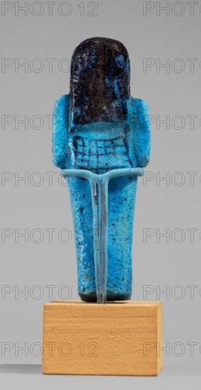 Queen Maatkare's shabti, Divine Wife of Amun, cartouches, hieroglyphs, uraeus iconographic, Maatkare ca. 1069–945 BC, wigs, funerary figurines, grave goods, mummy-shaped figurine. Arms crossed on the chest, the right folded over the left. Hoes in each hand. On the back is a basket slung on strings over both shoulders. Facial details, wig, and tools painted in a dark color. On the head is a three-part smooth wig. A uraeus above the forehead. Bracelets on the wrists. Painted inscription on the front of the legs in one wide column framed with lines: Shd Osiris, the wife of the god, the lord of both lands, Maat-ka-re. Third Intermediate Period; 21st Dynasty, Egypt; shabti; height 12 cm, width 4.4 cm., A small standing figurine displayed on a rectangular wooden block, with a glossy bright blue body showing mottled texture and darker blue and black areas on the head and upper torso; the figure has a smooth rounded head that is predominantly dark with patches of blue, short stubby arms at its...