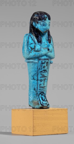 Queen Maatkare's shabti, Divine Wife of Amun, cartouches, hieroglyphs, uraeus iconographic, Maatkare ca. 1069–945 BC, wigs, funerary figurine, grave good, mummy-shaped figurine. Arms crossed on the chest, right folded over left, holding hoes in each hand. On the back is a basket slung on strings over both shoulders. Facial details, wig, and tools painted in a dark color. Three-part smooth wig; a uraeus above the forehead; bracelets on the wrists. Painted inscription on the front of the legs in one wide column framed with lines: Shd Osiris, the wife of the god, the lord of both lands, Maat-ka-re. Third Intermediate Period; 21st Dynasty, Egypt. Shabti. Height 12 cm, width 4.4 cm., A small standing figurine with crossed arms mounted on a rectangular block; the figurine is predominantly bright turquoise with darker blue and black areas on the hair, face, and details, showing a slightly worn or mottled surface; the figure wears a long garment that extends to the base and has darker linear m...