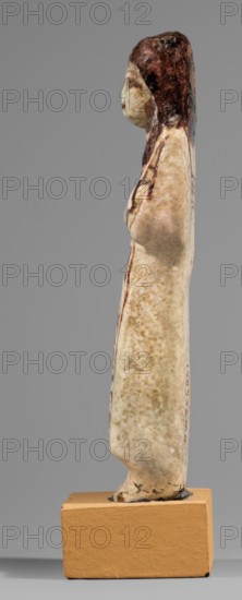 Ushabti of Mrs. As, hieroglyphs, As ca. 1295–1069 BC, usekh necklace, hoes, wigs, grave goods, figurine in living costume, loincloth reaching to the ankles. Arms crossed on the chest, right folded over left. Hoes in both hands, basket on the back slung over both shoulders. A usekh necklace on the chest, wide bracelets on the wrists. On the head is a smooth, double wig. Round face, poorly defined nose, narrow mouth, large eyes. The wig, eyes, eyebrows, mouth, necklace, bracelets, hoes, and bag are painted in brown with a burgundy tint. The inscription is painted and runs in one vertical line on the front of the headband: Shd Osiris, Lady of the House of As. Malgorzata Korzeniowska, c. 1295–c. 1069 BC; New Kingdom, 19th–20th Dynasty, Egypt, shabti; height 13.2 cm, width 4.5 cm, A slender vertical figure carved in a pale cream material stands on a rectangular tan base; the figure has darker brown coloring concentrated on the head and upper back, with mottled beige and light brown areas al...