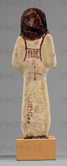 Ushabti of Mrs. As, hieroglyphs, As ca. 1295–1069 BC, usekh necklace, hoes, wigs, grave goods, Figurine in living costume, loincloth reaching to the ankles. Arms crossed on the chest, right folded over left. Hoes in both hands, basket on the back slung over both shoulders. A usekh necklace on the chest, wide bracelets on the wrists. On the head is a smooth, double wig. Round face, poorly defined nose, narrow mouth, large eyes. The wig, eyes, eyebrows, mouth, necklace, bracelets, hoes, and bag are painted brown with a burgundy tint. The inscription is painted and runs in one vertical line on the front of the headband reading: Shd Osiris, Lady of the House of As. Malgorzata Korzeniowska, c. 1295–c. 1069 BC; New Kingdom, 19th–20th Dynasty, Egypt, shabti; height 13.2 cm, width 4.5 cm, A small carved figure stands on a rectangular base, with an elongated pale body showing a mottled off-white and beige surface and a few darker speckles, topped by a rounded head that is darker brown with area...
