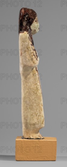 Ushabti of Mrs. As, hieroglyphs, As ca. 1295–1069 BC. Figurine in living costume, loincloth reaching to the ankles. Arms crossed on the chest, right folded over left. Hoes in both hands, basket on the back slung over both shoulders. Usekh necklace on the chest, wide bracelets on the wrists. Smooth, double wig; round face, poorly defined nose, narrow mouth, large eyes. The wig, eyes, eyebrows, mouth, necklace, bracelets, hoes, and bag are painted in brown with a burgundy tint. The inscription is painted, running in one vertical line on the front of the headband: Shd Osiris, Lady of the House of As. c. 1295–1069 BC; New Kingdom, 19th–20th Dynasty, Egypt. Shabti; height 13.2 cm, width 4.5 cm., A narrow upright figurine photographed in profile standing on a rectangular base, showing a smooth, elongated body with a rounded head and a suggestion of an arm along the torso; the object displays a palette of cream and beige tones across the body with darker brown concentrated on the top of the h...