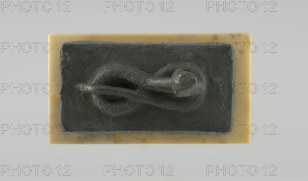 Snake sarcophagus, snakes; possibly representing the cobra Renenutet may also be Atum; reliquary-sarcophagus with snake with raised head coiled on a rectangular sarcophagus; Late Period, Egypt, 664–332 BC; figurine; height 3.3 cm, width 5.5 cm, depth 5 cm., A rectangular dark greenish object with a rough textured surface rests on a smooth rectangular base that is a pale yellow with slightly darker marbling; on the top of the dark object a sculpted form resembling a coiled, sinuous shape with a rounded thicker end sits, showing subtle surface patterns and variations in green and darker tones, and the background is a light neutral gray.