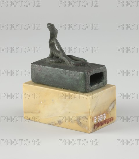 Snake sarcophagus; Snake with raised head, coils on a rectangular reliquary-sarcophagus—probably the cobra Renenutet, although it may also be Atum. Late Period, Egypt, 664–332 BC. Earthenware figurine reliquary/sarcophagus. Height 3.3 cm; width 5.5 cm; depth 5 cm., A small dark greenish object with a rough, weathered surface sits atop a yellow-beige rectangular block of stone with lighter and darker marbling; the green object has an irregular hollow opening on one side and a raised, elongated form on top that curves upward, with visible texture and slight variations of darker and lighter green.