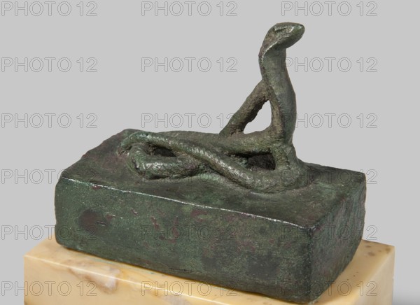 Snake sarcophagus, snakes, Atum mythology, Renenutet mythology, reliquaries, sacred animals. Scen.Gal.Eg. Snake with raised head, coils on a rectangular reliquary-sarcophagus. This is probably the cobra Renenutet, although it may also be Atum. 664–332 BC; Late Period, Egypt. Figurine; height 3.3 cm, width 5.5 cm, depth 5 cm., A small bronze sculpture of a coiled snake mounted on a rectangular bronze base, showing the snake's head raised and neck curved, with a textured surface and visible patina in shades of green and darker green, sitting on a light beige marble plinth with subtle veining, all set against a plain pale gray background.