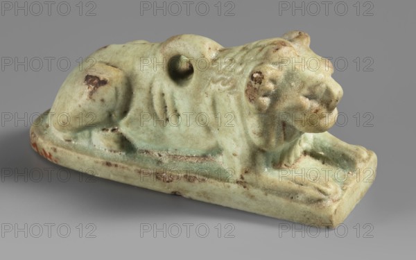 Amulet in the shape of a lion lying on a flat base, with paws stretched forward, head facing straight ahead, and a loop for hanging on its back. The ribs and mane are schematically marked with diagonal engraved lines. Amulet; material unspecified. Late Period, Egypt, 664–332 BC. Height 2.6 cm; width 5.2 cm., A small sculpted figure of a reclining animal on a rectangular base, showing a rounded body, a pronounced head with a textured mane, front legs extended forward, and a curled tail area with a hole through the back; the surface appears worn with patches of darker discoloration and rough spots, and the predominant color is a pale green with areas of brown and tan weathering set against a neutral gray background.
