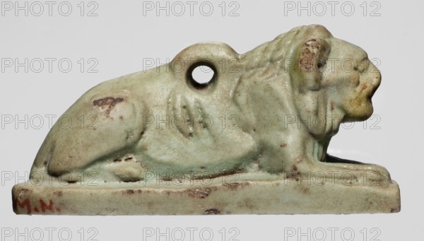 Amulet in the shape of a lion lying on a flat base, with paws stretched forward, head facing straight ahead, and a loop for hanging on its back. The ribs and mane are schematically marked with diagonal engraved lines. Amulet; material unspecified. Height 2.6 cm, width 5.2 cm. Late Period, Egypt, 664–332 BC., A small sculpted animal figure shown in profile, lying atop a rectangular base with a rounded body, pronounced mane, and a circular hole through its upper back; the surface shows a worn, slightly pitted texture with areas of discoloration and small chips. The visible colors include pale green, muted beige, light brown, and hints of darker brown in worn spots, with a small area of reddish-brown on the base.