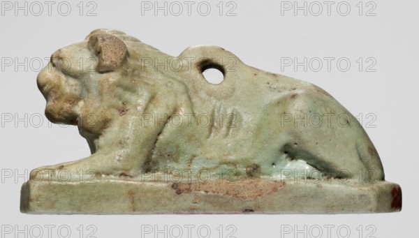 Amulet in the shape of a lion lying on a flat base, with paws stretched forward, head facing straight ahead, and a loop for hanging on its back. The ribs and mane are schematically marked with diagonal engraved lines. Amulet lion, Late Period, Egypt, 664–332 BC. Height 2.6 cm; width 5.2 cm., A small sculpted figure of a reclining animal on a rectangular base, shown in profile facing left, with a rounded body, visible head, front paw resting on the base, and a raised hind area; the surface shows a smooth, worn texture with a few rough spots and pitting, and there is a hole through the upper midsection of the figure; the visible colors are pale green, light brown, tan, and small areas of darker brown and faint reddish tones.