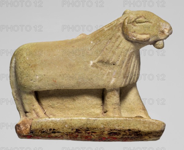 Amulet in the shape of a ram standing on a stand with curled horns and a short beard; the fur around the head is marked with simple engraved lines. Amulet; height 2.5 cm, width 4 cm, depth 1.1 cm. Late Period, Egypt, 664–332 BC., A small sculpted animal figure in shades of pale green and yellow with areas of darker greenish speckling, showing a rounded body, a mane suggested by incised vertical lines, a head with a short snout and small rounded ears, and a tongue or curved projection at the mouth; it stands on a rectangular base that is similarly colored with patches of darker brown and small traces of red along the lower edge, and the surface shows weathered texture and small pits.