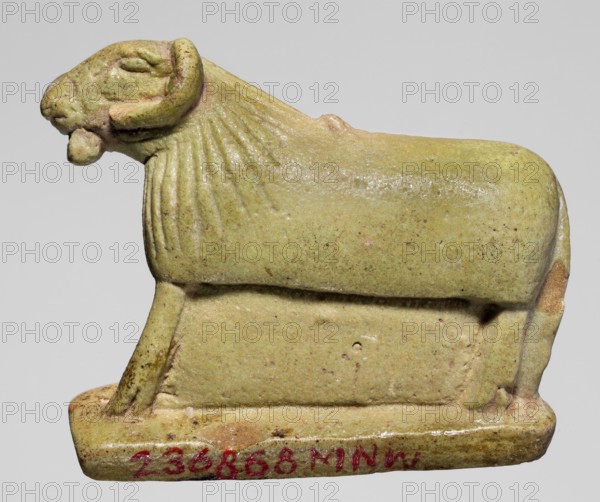 Amulet in the shape of a ram standing on a stand with curled horns and a short beard. The fur around the head is marked with simple engraved lines. Amulet; height 2.5 cm, width 4 cm, depth 1.1 cm. Late Period, Egypt, 664–332 BC., A small sculpted figure of a ram or sheep rendered in a smooth stone-like material, shown in profile facing left with a rounded body and stylized mane lines along the neck; the head displays curved horns and a protruding lower jaw. The figure rests on a rectangular base with slightly beveled edges. The visible colors are pale green, yellowish-green, light tan, with small darker speckles and a few areas of brownish discoloration.