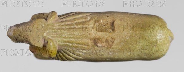 Amulet in the shape of a ram standing on a stand with curled horns and a short beard. The fur around the head is marked with simple engraved lines. Amulet; material unspecified. Late Period, Egypt, 664–332 BC. Dimensions: height 2.5 cm, width 4 cm, depth 1.1 cm., A close-up of a small carved object shaped like an elongated animal form, shown horizontally against a pale gray background; the object has a mottled surface in shades of olive green, yellow-green, brown, and a little tan, with areas of slightly glossy wear and small darker speckles; one end tapers into a rounded snout with a pale worn tip, and a protruding rounded ear-like shape near the front; the body shows incised linear grooves radiating from the head area and a few shallow rectangular depressions on the upper surface, with subtle textural pitting and smoother rounded contours toward the rear.