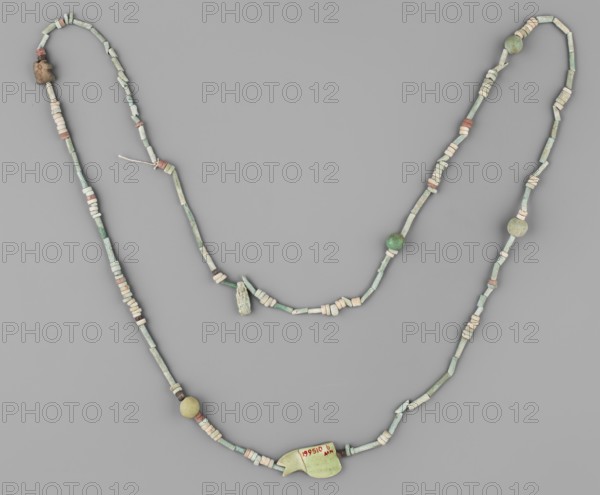 Necklace, repossession Radom. Egyptian deities, udjat eyes, beads, spherical beads, ornaments. Necklace consisting of 67 straw-shaped beads, 117 flat rings, 10 double flat rings with two holes, 4 balls, 2 udjat eyes 1 eyelet in 2 parts, 1 deity figurine without preserved feet. Predominant color: green; single beads brick-red, brown, and white. 664–332 BC, Late Period, Egypt. Amulet, jewelry; height 98 cm., A beaded necklace arranged on a flat gray background, composed of a string of small cylindrical and disc-shaped beads in pale green and off-white, with several beads in muted pink and some round beads in green and yellowish tones; a larger pale green pendant with an irregular shape is attached near the lower center, and there are a few small rough stone or clay beads in beige and brown along the strand.