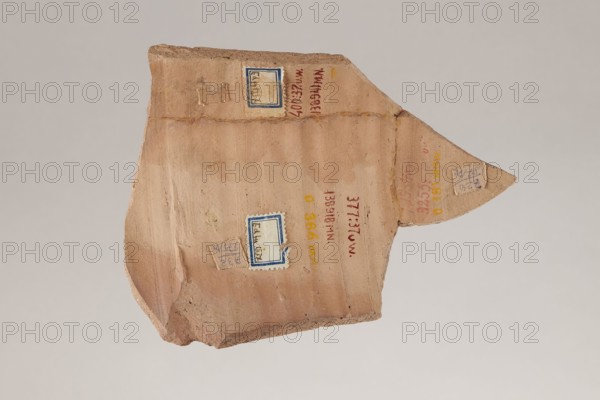 Greek ostraka – two tax receipts, taxes, receipts. Fragment of a clay vessel with two tax receipts paid by Panibekis, son of Achillas Rufus, 27/04/161, Edfu Apollonopolis Wielka. Ostraka; height 11.5 cm, width 15.5 cm, depth 0.5 cm, weight 0.136 kg., A fragment of a flat, irregularly shaped pottery shard with a triangular projection, showing a surface of muted tan and light brown clay with subtle horizontal striations and areas of slightly darker brown; a few small rectangular paper or fabric labels are affixed, outlined in blue and beige, with one label on the triangular tip and another closer to the center, and there are a few small reddish and yellow marks on the clay surface; the shard has rough, broken edges showing a gritty, darker brown interior and a thin rim along one side, all set against a pale gray-beige background.