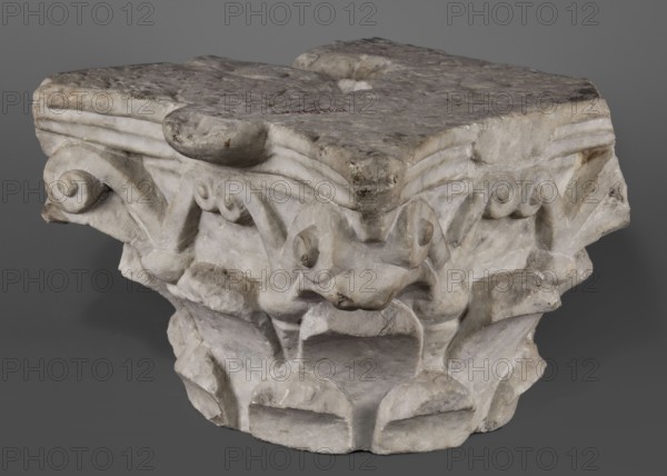Corinthian capital, Corinthian capital composed of a calathos and thick stems that bifurcate and terminate in volutes supporting the abacus. The crown of leaves, formed by two rows of eight full leaves, occupies half the height of the capital. The leaves are rounded and angled outward. Thick caules with lacings in the form of thick rings grow from the upper row. The volute stems are in the form of flat ribbons. The outer volutes are significantly larger than the inner ones, meeting directly under the projection of the abacus rosette. The abacus is profiled, two-part with extended corners. Two corners, including the volutes, are upholstered. The edges of the leaves are chipped. 4th–5th century late antique period, Roman Empire; architectural element / capital; height 24 cm, width 34 cm, depth 33 cm, weight 42.5 kg., A stone carving fragment with a weathered surface in shades of white, gray, and beige, showing carved volute-like spiral forms and rounded protrusions; the top appears flat ...