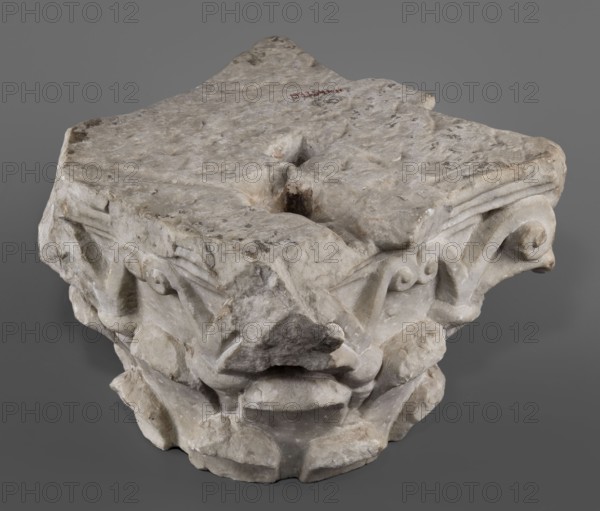 Corinthian capital, Corinthian capital composed of a calathos and thick stems that bifurcate and terminate in volutes supporting the abacus. The crown of leaves, formed by two rows of eight full leaves, occupies half the height of the capital. The leaves are rounded and angled outward. Thick caules with lacings in the form of thick rings grow from the upper row. The volute stems are in the form of flat ribbons. The outer volutes are significantly larger than the inner ones, meeting directly under the projection of the abacus rosette. The abacus is profiled, two-part with extended corners. Two corners, including the volutes, are upholstered. The edges of the leaves are chipped. 4th–5th century late antique period, Roman Empire; architectural element / capital; materials: Roman architecture ornamentation with acanthus leaves; height 24 cm, width 34 cm, depth 33 cm, weight 42.5 kg., An off-white carved stone fragment with areas of gray and beige discoloration sits against a neutral gray b...