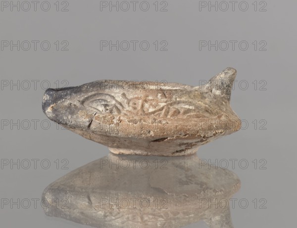 Oil lamp, deposit provenance, Islamic art, Arabic culture, functional ceramics, Arabic pottery, oil lamp depicting everyday life in the Middle East; plant tendril decoration, traces of use. Oval lamp with short burner channel surrounded by a shaft similar to the disc. Large oil hole occupying the entire surface of the disc. Arms decorated with plant tendrils. Conical handle. Ring-shaped base with a star in the center. Type A oil lamp according to the typology of Kubiak of Fustat 9th–11th century; similar lamps have been found in Umayyad contexts late 7th–8th century. 2nd half 7th–8th century, Syro-Palestine; oil lamp, height 3.3 cm, width 11.0 cm, depth 9.8 cm., An aged shallow vessel with a rounded base and a narrow spout sits against a plain gray background; the object shows a weathered surface with a mix of beige, tan, brown, gray, and darker charcoal tones, with worn textured areas, chips, and rough patches, and a reflected image of the vessel below creating muted mirrored beige an...