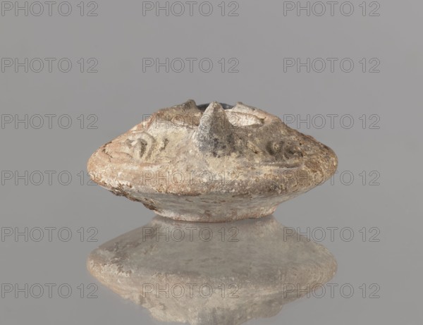 Oil lamp, deposit provenance, Islamic art, Arabic culture, functional ceramics, Arabic pottery, oil lamp used in everyday life in the Middle East. Oval lamp with short burner channel surrounded by a shaft like a disc; large oil hole occupying the entire surface of the disc. Arms decorated with plant tendrils. Conical handle. Ring-shaped base with a star in the center. Type A oil lamp according to Kubiak's typology of Fustat 9th–11th century; similar lamps have been found in Umayyad contexts late 7th–8th century. 2nd half 7th–8th century, Syro-Palestine; oil lamp; height 3.3 cm, width 11.0 cm, depth 9.8 cm., A weathered round object with a raised central cone sits on a reflective surface, showing a mirrored underside; the object has an irregular textured surface with rough edges and carved or eroded shallow depressions, and its colors include beige, tan, brown, gray, and off-white.