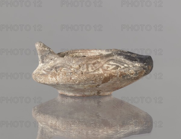 Oil lamp, Islamic art; Arabic culture; functional ceramic oil lamp. Oval lamp with short burner channel surrounded by a shaft-like disc; large oil hole occupying the entire surface of the disc. Arms decorated with plant tendrils; conical handle; ring-shaped base with a star in the center. Type A oil lamp Kubiak typology of Fustat, though similar lamps appear in Umayyad contexts late 7th–8th century. 2nd half 7th–8th century, Syro-Palestine. Height 3.3 cm; width 11.0 cm; depth 9.8 cm. Traces of use., An aged shallow bowl or lamp-like ceramic object sits on a rounded pedestal, showing worn, textured surfaces with rough edges and chips; the rim has a small pointed spout on one side and an irregular darkened area on the opposite side, with carved or incised decorative motifs around the upper body. The object displays a palette of muted earthy colors including beige, tan, brown, gray, and areas of darker charcoal, with lighter off-white encrustations and subtle mottling across its surface; ...