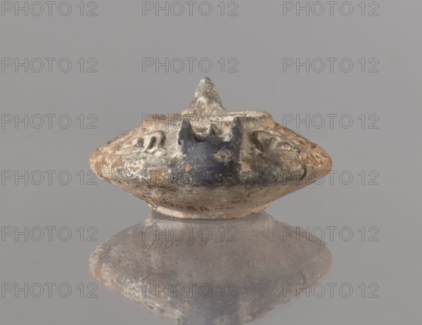 Oil lamp, deposit provenance, Islamic art, Arabic culture, functional ceramics, Arabic pottery, oil lamp used in everyday life in the Middle East; oval lamp with short burner channel surrounded by a shaft similar to the disc. Large oil hole occupying the entire surface of the disc. Arms decorated with plant tendrils. Conical handle. Ring-shaped base with a star in the center. Type A oil lamp according to the typology of Kubiak of Fustat 9th–11th century; similar lamps have been found in Umayyad contexts late 7th–8th century. 2nd half 7th–8th century, Syro-Palestine; height 3.3 cm, width 11.0 cm, depth 9.8 cm., An aged, rounded object with a textured, worn surface sits against a solid gray background and shows a mix of colors including beige, brown, dark gray, and hints of black; the object has a raised central point and irregular indentations and grooves around its upper edge, with patches of lighter and darker areas creating a mottled appearance, and the lower portion tapers slightly ...