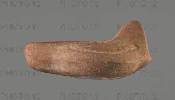Oil lamp, Horse iconography. Lamp pressed from a mold. Light red clay with brownish-red engobe. Almond-shaped reservoir. Ring-shaped, slightly concave base with a mold mark of two notches letter lambda or simplified alpha. Single groove runs from the base toward the nozzle. Solid nozzle. Handles decorated with a simplified winding vine s-scroll motif. Slightly concave discus with two olive holes, decorated with a depiction of a horse galloping left. Medium-length burner with an oval end and a round wick hole. No traces of use. 4th–5th century, Tunisia. Oil lamp; height 10.7 cm, width 6.9 cm., A single irregularly shaped object with a textured, rough surface in shades of reddish-brown, terracotta, and hints of pale beige set against a flat gray background; the object has a rounded elongated body with a raised, curved ridge on one side and a taller, sculpted protrusion at one end, showing variations in surface texture and subtle areas of lighter and darker coloration.