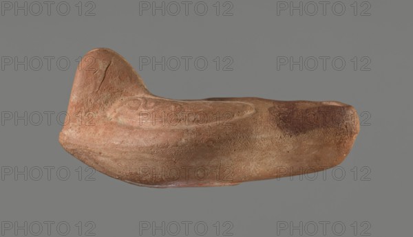 Oil lamp, Horse iconography. Lamp pressed from a mold. Light red clay with brownish-red engobe. Almond-shaped container. Ring-shaped, slightly concave base with a mold mark of two notches letter lambda or simplified alpha. A single groove runs from the base toward the nozzle. Solid nozzle. Arms decorated with a simplified winding vine s-scroll motif. Slightly concave discus with two olive holes, decorated with a depiction of a horse galloping left. Medium-length burner with an oval end and a round wick hole. No traces of use. 4th–5th century, Tunisia. Oil lamp. Height 10.7 cm, width 6.9 cm., A single worn, boat-shaped clay object is shown against a plain gray background; the object has an irregular rounded form with a higher curved end and a lower tapered end, a textured surface with visible scratches and pits, and a shallow, oval recessed area on its top. The colors visible include reddish-brown, terracotta, rust, beige, and small areas of darker brown, with subtle lighter patches and...