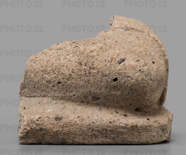 Votive offering in the form of an elbow, 4th–3rd century BC, Etruria; sculpture, votive offering; height 7.5 cm, width 10 cm, A weathered stone object with a rough, porous surface sits against a plain gray background; the stone shows a tan to light brown color with darker brown and black speckles and small pits, and it rests on a rectangular base that has a slightly lighter tan tone along its front edge, while the upper portion has a rounded, irregular form with subtle indentations and a few darker flecks.