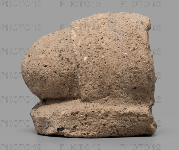 Votive offering in the form of an elbow, 4th–3rd century BC, Etruria; sculpture, votive offering; height 7.5 cm, width 10 cm, A roughly textured, irregularly shaped stone object sits against a plain gray background, showing a weathered surface with pitting and embedded small fragments; the stone appears in shades of light brown and tan with darker speckles of black and gray and a few tiny white flecks, and the base rests on a smooth, slightly darker gray surface.