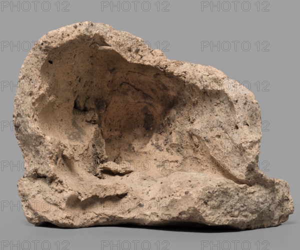 Votive offering in the form of an elbow, 4th–3rd century BC, Etruria; sculpture; votive offering; height 7.5 cm, width 10 cm, A roughly shaped cavity of porous beige and tan rock sits against a flat gray background, showing irregular rough surfaces, pitted textures, and small dark speckles; the interior of the cavity appears darker with brownish shading and varied erosion patterns, while the exterior edges reveal lighter sandy tones and crumbly contours with subtle shadows cast beneath and around the base.