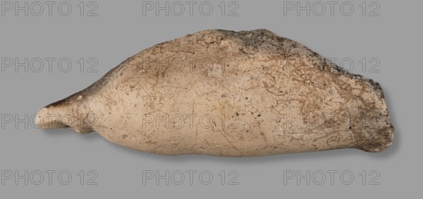 Votive offering in the form of a phallus, votive offering, phalluses, 332–30 BC; Hellenistic period, Etruria, Caere, height 6 cm, width 15 cm, depth 6 cm, weight 0.21 kg, length 15.3 cm, A horizontally oriented, elongated ceramic vessel with a tapered end and a wider rounded opposite end rests against a neutral gray background; the vessel surface shows a beige to light tan base color with mottled areas of darker brown and gray discoloration, scattered small dark speckles across its body, and faint linear scratches and marks etched into the surface; the tapered end appears slightly darker with a shadowed underside, while the wider end has a rougher, more irregular texture and darker brown-gray crusting.