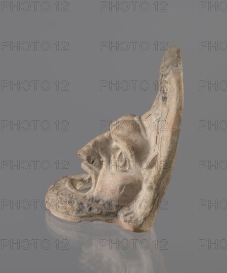 Tragic mask, terracotta, 4th–3rd century BC, Southern Italy. Tragic mask depicting a male face, likely a decorative element antefix?. The lower part with the beard underside edges flat is set at a near-perpendicular angle to the facial part and forms the base. Realistic theatrical features: protruding cheekbones, deep furrows around mouth and eyes; mouth wide open with nearly horizontal lower lip and arched upper lip; large, deep-set eyeballs with circular cut-out holes at slightly different heights right higher than left; slightly hooked nose with horizontal wrinkles; thick eyebrows, vertical forehead wrinkles, and a convex forehead. Above the forehead is a flat, high onkos with schematically marked locks of hair, bounded at top and sides by folds of cloth. Light orange clay, unevenly fired at the fracture turning red. Traces of polychrome—red, on the cheek, black, on the beard and hair. The upper right part above the forehead has been reconstructed., A sculpted fragment of a face in ...