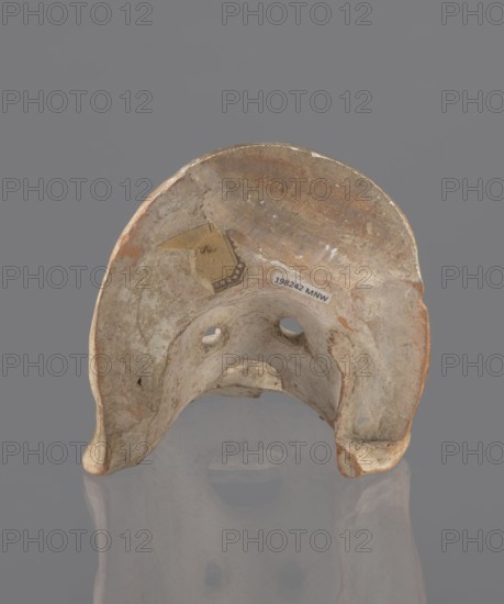 Tragic mask, terracotta, 4th/3rd century BC, Southern Italy. Tragic mask depicting a male face, likely a decorative element antefix?. Lower part with beard underside edges flat set at near-perpendicular angle to facial part forming the base. Realistic representation of a theater mask: protruding cheekbones, deep furrows around mouth and eyes; mouth wide open with lower lip almost horizontal and upper lip arched. Large, deep-set eyeballs with circular holes cut out, positioned at slightly different heights right higher than left. Nose slightly hooked with horizontal wrinkles; thick eyebrows; vertical wrinkles in center of forehead; convex forehead. Above the forehead a flat, high onkos with schematically marked locks of hair, bounded at top and sides by folds of cloth. Light orange clay, unevenly fired at fracture turning red. Traces of polychrome—red paint on cheek, black on beard and hair. Upper right part above the forehead has been reconstructed. mask, A weathered curved object with...