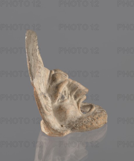 Tragic mask, terracotta, 4th/3rd century BC, Southern Italy. Tragic mask depicting a male face, likely a decorative element antefix?. The lower part with the beard underside edges flat is set at a near-perpendicular angle to the facial part and forms the base. Realistic theatrical features: protruding cheekbones; deep furrows around mouth and eyes; wide open mouth with almost horizontal lower lip and arched upper lip; large deep-set eyeballs with circular cut-out holes positioned at slightly different heights right higher than left; slightly hooked nose with horizontal wrinkles; thick eyebrows; vertical central forehead wrinkles; convex forehead. Above the forehead is a flat, high onkos with schematically marked locks of hair, bounded at top and sides by folds of cloth. Light orange clay, unevenly fired at the fracture turning red. Traces of polychrome—red, on cheek, black, on beard and hair. The upper right part above the forehead has been reconstructed., A weathered sculpted fragment...