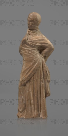 Terracotta figurine of a veiled woman, Dancers, molded female figurine, Tanagra figurine, terracotta, fine-grained red clay. Standing woman in contrapposto. Dressed in a long chiton and himation covering the head, lower face, and upper torso. Both arms bent: right hand placed in front on the breasts, left behind on the back. Probably a figure of a masked dancer. Condition: base stamped, head glued. 3rd century BC, Tanagra Greece. Sculpture. Height 15.9 cm, width 4.8 cm., A single standing figure sculpted from a brown material against a gray background, wearing a draped garment that wraps across the torso and falls in long vertical folds, with one arm bent at the waist creating a gathered area of fabric at the hip and the other arm less defined, the head covered by a close-fitting hood that obscures facial details, surface showing slight irregularities and worn areas, and visible colors including shades of brown on the figure and solid gray in the background.