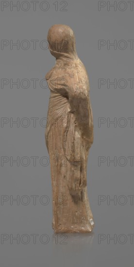 Terracotta figurine of a veiled woman, Dancers, molded female figurine in the Tanagra style, terracotta, fine-grained red clay. Figure of a standing woman in contrapposto. She wears a long chiton and a himation covering the head, lower face, and upper torso. Both arms are bent — right hand placed in front on the breasts, left behind on the back. Probably a masked dancer. Condition: base stamped, head glued. 3rd century BC, Tanagra Greece. Sculpture. Height 15.9 cm, width 4.8 cm., A worn statue of a standing figure draped in long flowing garments, shown in profile and facing left, with a smooth rounded head and a wrapped cloth over the head and shoulders; the surface shows textured weathering and small chips, and the base is flat and reflective. The visible colors are shades of brown, tan, beige, gray, and a muted clay tone, set against a solid mid-gray background.