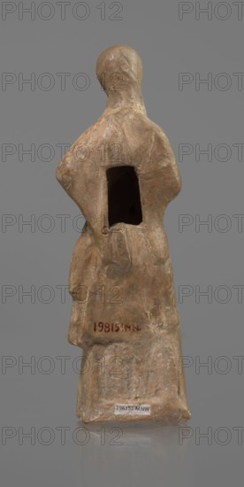Terracotta figurine of a veiled woman, dancer, molded female figurine in fine-grained red clay Tanagra figurine. Figure of a standing woman in contrapposto. She wears a long chiton and himation covering the head, lower face, and upper torso. Both arms are bent — right hand placed in front on the breasts, left behind on the back. Probably the figure of a masked dancer. Condition: base stamped, head glued. Date: 3rd century BC. Place of origin: Tanagra Greece. Medium: terracotta sculpture. Dimensions: height 15.9 cm, width 4.8 cm., A standing figurine made of a rough textured material in shades of tan, beige, and light brown, shown from the back with a rounded head and simplified body forms; the figure has a rectangular hollow in the upper back and subtle carved folds suggesting clothing, with areas of lighter and darker tan tones and small patches of pale off-white on the surface, set against a plain neutral gray background.