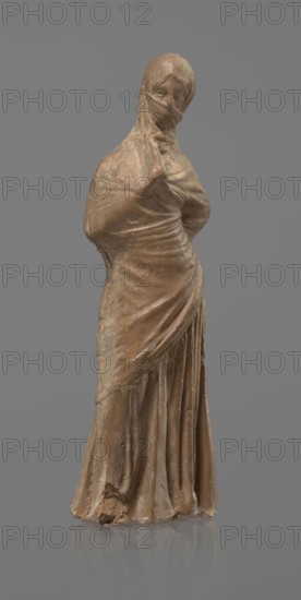 Terracotta figurine of a veiled woman, dancer, molded Tanagra figurine in fine-grained red clay. Figure of a standing woman in contrapposto wearing a long chiton and himation covering the head, lower face, and upper torso. Both arms bent — right hand placed in front on the breasts, left behind on the back. Probably a masked dancer. Condition: base stamped, head glued. 3rd century BC, Tanagra Greece. Sculpture. Height 15.9 cm, width 4.8 cm., A standing sculpture of a draped figure shown against a plain gray background, the figure appears wrapped in a long flowing garment that covers the head and body, with visible folds and creases in the fabric and a wrapped section across the lower face; the surface shows an earthy brown color with variations of lighter beige and subtle worn spots, and there are areas of darker brown patina and tiny surface imperfections on the dress and base.
