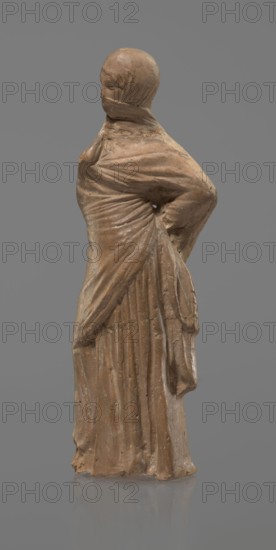 Terracotta figurine of a veiled woman, Dancers, molded female figurine in the Tanagra style, fine-grained red clay. Figure of a standing woman in contrapposto wearing a long chiton and a himation covering the head, lower face, and upper torso. Both arms bent — right hand placed in front on the breasts, left behind on the back. Probably the figure of a masked dancer. Condition: base stamped, head glued. 3rd century BC, Tanagra Greece. Sculpture; height 15.9 cm, width 4.8 cm., A standing statue of a robed figure rendered in a brownish material against a plain gray background, showing draped clothing with a hooded head and covered face, visible folds and pleats in the garment, a posture with one arm bent behind the back creating a gathers at the waist, textured surface with worn and slightly uneven areas, and subtle variations of brown and tan tones contrasted with the neutral gray backdrop.