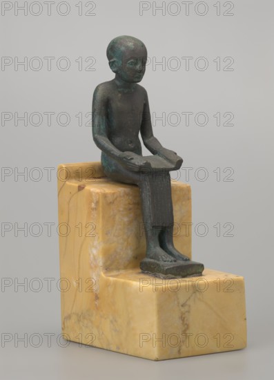 Figurine of Imhotep, Imhotep ca. 2667–2648 BC; mythology. Figurine of a seated Imhotep holding an unrolled papyrus on his lap; the palms of both hands hold the ends of the papyrus. Materials: papyri. Subject: Egyptian deities; male figurines. Date: 664–332 BC, Late Period, Egypt. Type: figurine. Dimensions: height 12 cm, width 3.2 cm., A small greenish bronze figure with a bluish patina sits on a stepped block of yellow and beige marbled stone, the figure seated with legs extended and hands holding a flat object across the lap, the figure's surface showing darker green and brown areas, the stone base displaying veins and mottled patterns in cream, beige, and pale yellow, all set against a plain light gray background.