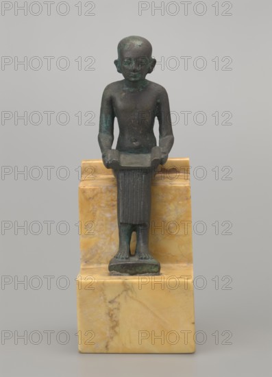 Figurine of Imhotep; seated male figure holding an unrolled papyrus on his lap, palms holding the papyrus ends. Imhotep ca. 2667–2648 BC; mythology. Materials: papyri? associated, Egyptian deities, male figurine. Date: 664–332 BC, Late Period, Egypt. Type: figurine. Dimensions: height 12 cm, width 3.2 cm., A small dark greenish bronze figure of a standing person with a smooth rounded head and simplified facial features, shown frontally with arms bent and hands holding a shallow rectangular object, wearing a vertically ribbed skirt that reaches the ankles and standing on a small rectangular base, all set on a polished yellow and cream marbled stone pedestal; the background is a uniform light gray.