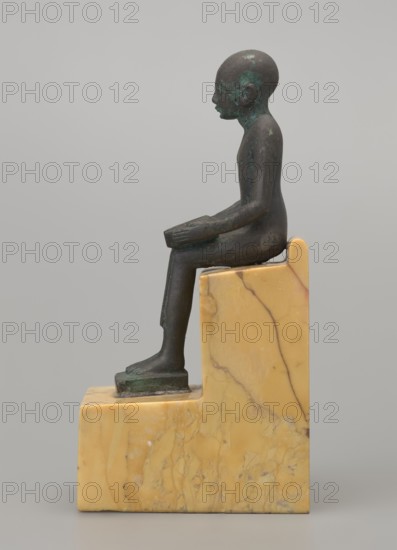 Figurine of Imhotep, Imhotep ca. 2667–2648 BC; mythology. Seated male figurine holding an unrolled papyrus on his lap, palms holding the ends of the papyrus. Material: papyri. Date: 664–332 BC, Late Period, Egypt. Height 12 cm, width 3.2 cm., A small dark bronze figurine of a seated person with a smooth rounded head and simplified facial features sits upright on a stepped block of yellowish marble with veining; the figure's arms rest on the thighs and the forearms extend forward, the legs bend at the knees with the feet placed on a lower ledge of the marble base, and patches of greenish patina are visible on parts of the bronze surface.