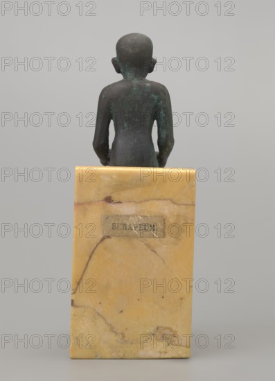 Figurine of Imhotep, Imhotep ca. 2667–2648 BC; papyri; Egyptian deities; male figurines. Figurine of a seated Imhotep holding an unrolled papyrus on his lap. The palms of both hands hold the ends of the papyrus. Date: 664–332 BC, Late Period, Egypt. Material: not specified; type: figurine; dimensions: height 12 cm, width 3.2 cm., A small dark greenish-brown seated figure viewed from the back, showing a rounded head with ears and a smooth back and arms, positioned on top of a rectangular yellow-beige marble base with natural veins and patches of darker tan and brown.