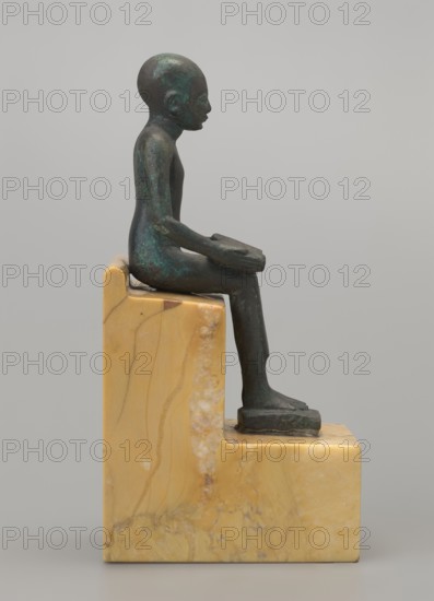 Figurine of Imhotep: seated Imhotep holding an unrolled papyrus on his lap, palms of both hands holding the ends of the papyrus. Inscribed/implied association with Imhotep ca. 2667–2648 BC; mythology. Materials: papyri depicted. Type: male figurine; style: Egyptian, Late Period. Date: 664–332 BC. Dimensions: height 12 cm, width 3.2 cm. Function: figurine., A small figurine of a seated person in a greenish dark bronze tone sits on a stepped block of yellowish beige marble with veins and patches of lighter and darker beige; the figure is shown in profile facing right with elongated limbs and a smooth rounded head, hands resting on the knees, feet placed on the lower step of the marble base, and the surface of the figure shows areas of darker and lighter greenish patina against the dark bronze.
