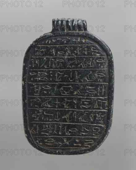 Heart scarab, Pectoral scarab with nine lines of hieroglyphic text containing Chapter 30B of the Book of the Dead; the chapter begins directly without the name of the owner. The scarab is fitted with a pendant, and the base is surrounded by a frame imitating a rope. ca. 1550–1295 BC; New Kingdom, 18th Dynasty, found at Antinoe. Scarab amulet; height 6 cm, width 4 cm, depth 2.2 cm., An oval, vertically oriented dark object with a rounded top and a small ridged projection at its upper edge, showing carved horizontal bands filled with repeated incised shapes; the surface is predominantly black with visible areas of brownish wear and a few greenish spots, and the background is a smooth neutral gray.