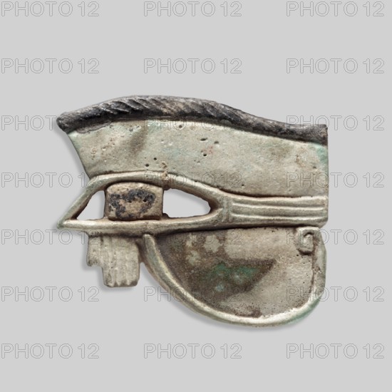 Amulet - Udjat Eye, Udjat eye designed as a flat plaque with details of the eye marked with a fairly wide engraved line. Eyebrow and pupil painted black. A pin placed above the eyebrow. ca. 747–656 BC; 25th Dynasty, Egypt, amulet, height 5.5 cm, width 7 cm, depth 1 cm, A carved object resembling a stylized eye shape with an upper brow-like ridge and a curved lower section, showing a worn, textured surface. The piece displays a palette of muted colors including beige, light gray, greenish patina, darker gray, brown, and small hints of reddish spots. The upper ridge has a darker gray, almost blackened appearance with a ridged texture, while the central eye area includes a recessed oval with mottled darker brown and gray. The lower curved section shows smoother wear with patches of greenish discoloration and lighter beige highlights, and there is a small protruding rectangular element beneath the eye opening. The background is a uniform light gray.