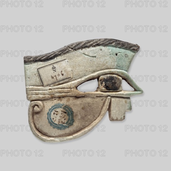 Amulet - Udjat Eye. Udjat eye designed as a flat plaque with details of the eye marked with a fairly wide engraved line. Eyebrow and pupil painted black. A pin placed above the eyebrow. Ca. 747–656 BC; 25th Dynasty, Egypt. Amulet; height 5.5 cm, width 7 cm, depth 1 cm., An object shaped like a stylized eye with a curved lower extension, made of a pale material with areas of light green and beige, and darker gray along the top edge; a rounded darkened central element sits within the eye opening, surrounded by lighter beige and green surfaces; there are a few worn and rough spots, a small hole near the top right edge, and a circular blue-green decorative mark on the lower curved portion; the piece shows signs of age with cracks and surface discoloration in colors including beige, light green, gray, dark brown, and blue-green.